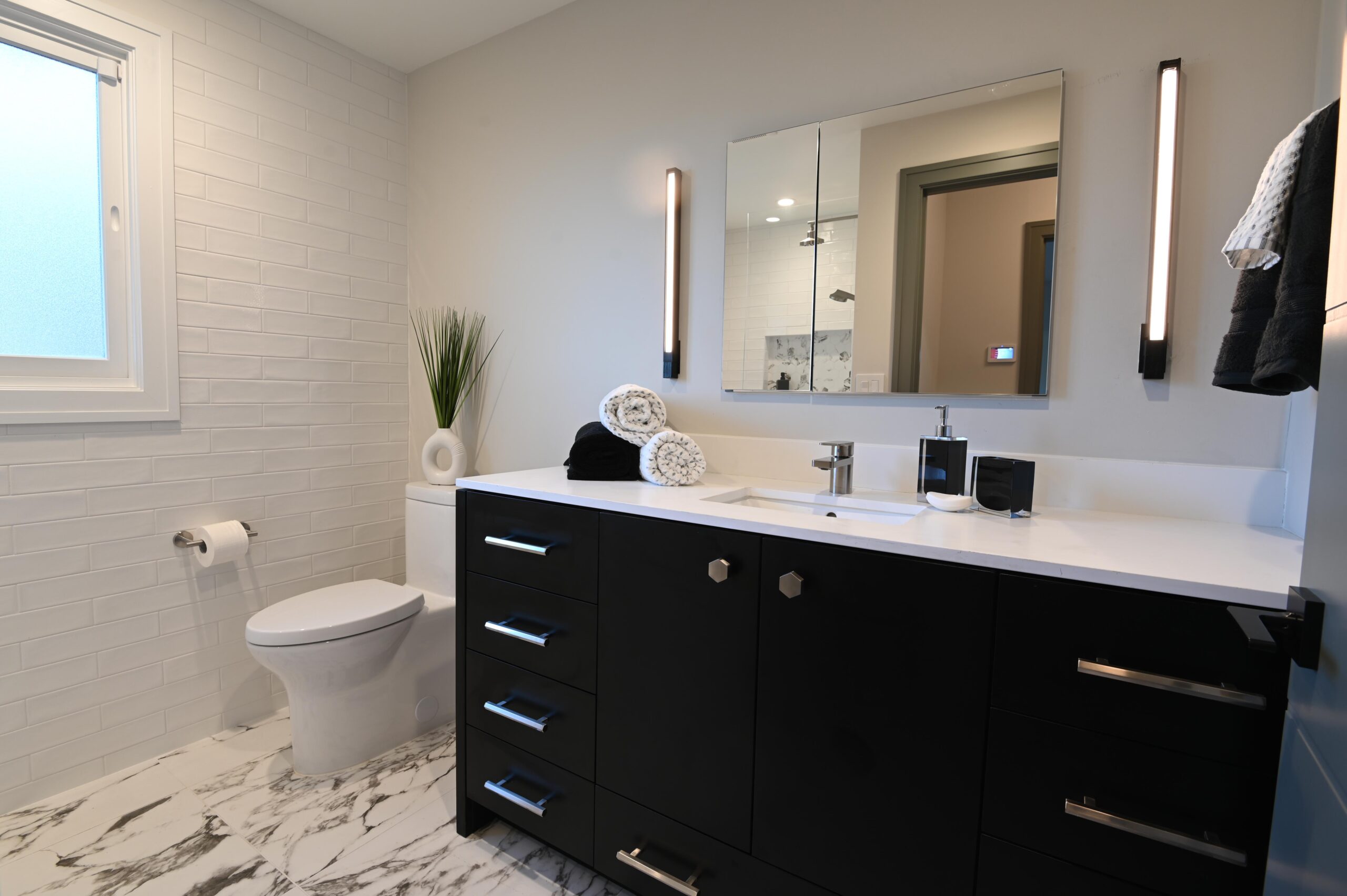 A modern bathroom features a black vanity with a white countertop, a wall-mounted mirror, a toilet, and a towel rack. Decorative items include rolled towels and a potted plant.